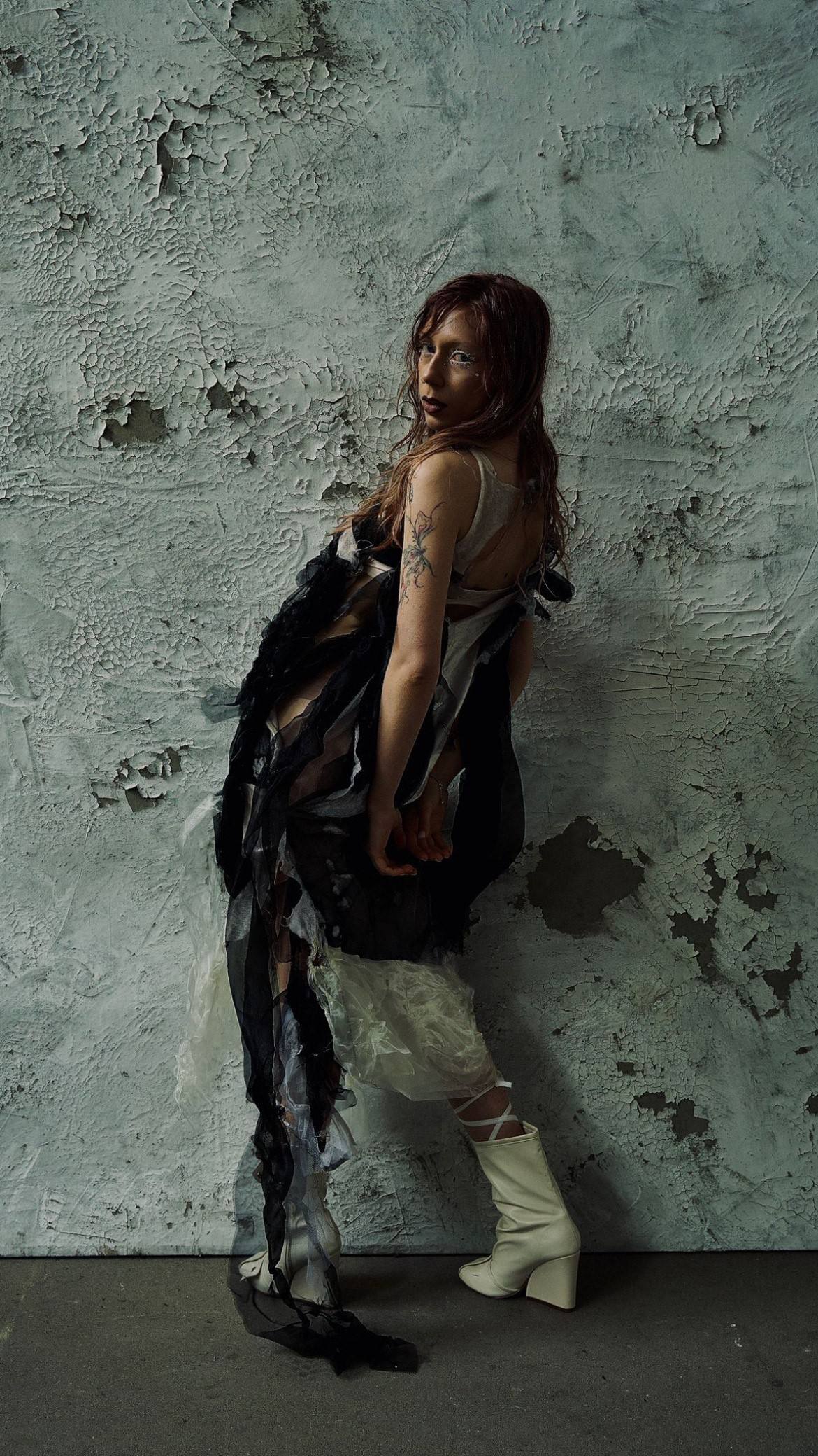Full-length portrait of a woman with long auburn hair in a dark, ragged layered dress with white ruffles and white boots, against a weathered teal wall