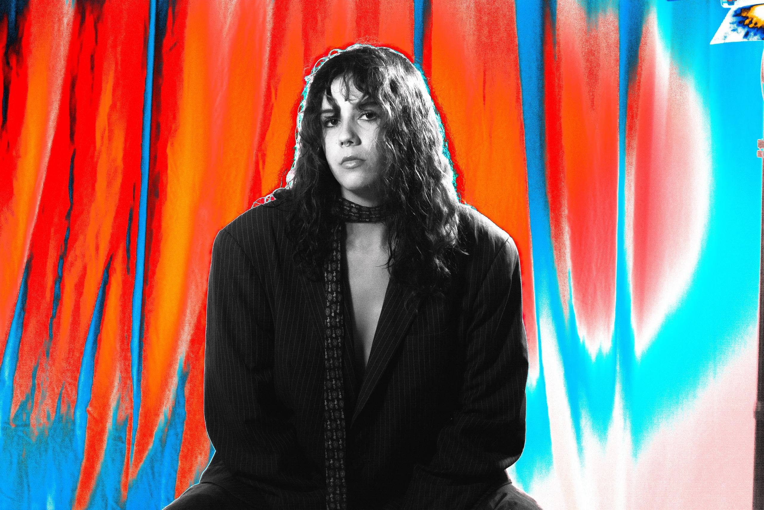 Black-and-white portrait of a woman with long dark curls, blazer and scarf, in front of vivid red and blue streaked curtains