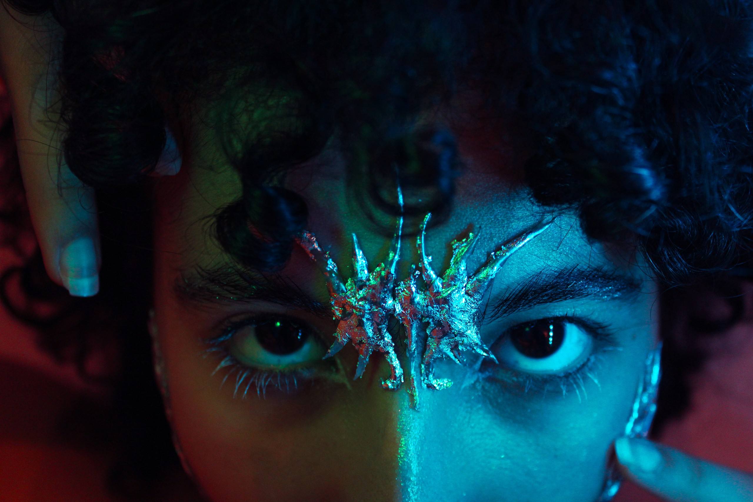Close-up of a face with a silver crystal growth on the nose and forehead, curly hair, split red and teal light