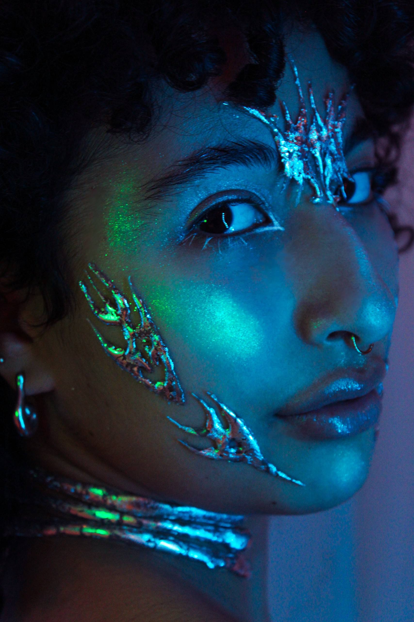 Portrait with chrome growths on the cheek and forehead and a chrome choker, in teal-blue light